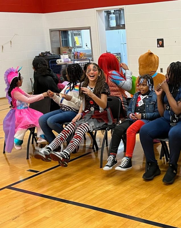 A group of girls wearing Halloween costumes sitting in a gymnasium, laughing.